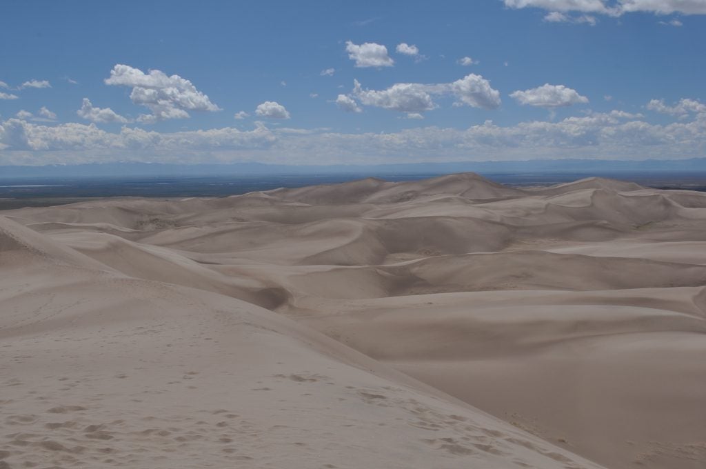 Expansive sand dunes with a cloudy, bright blue sky and mountain peaks on the horizon.